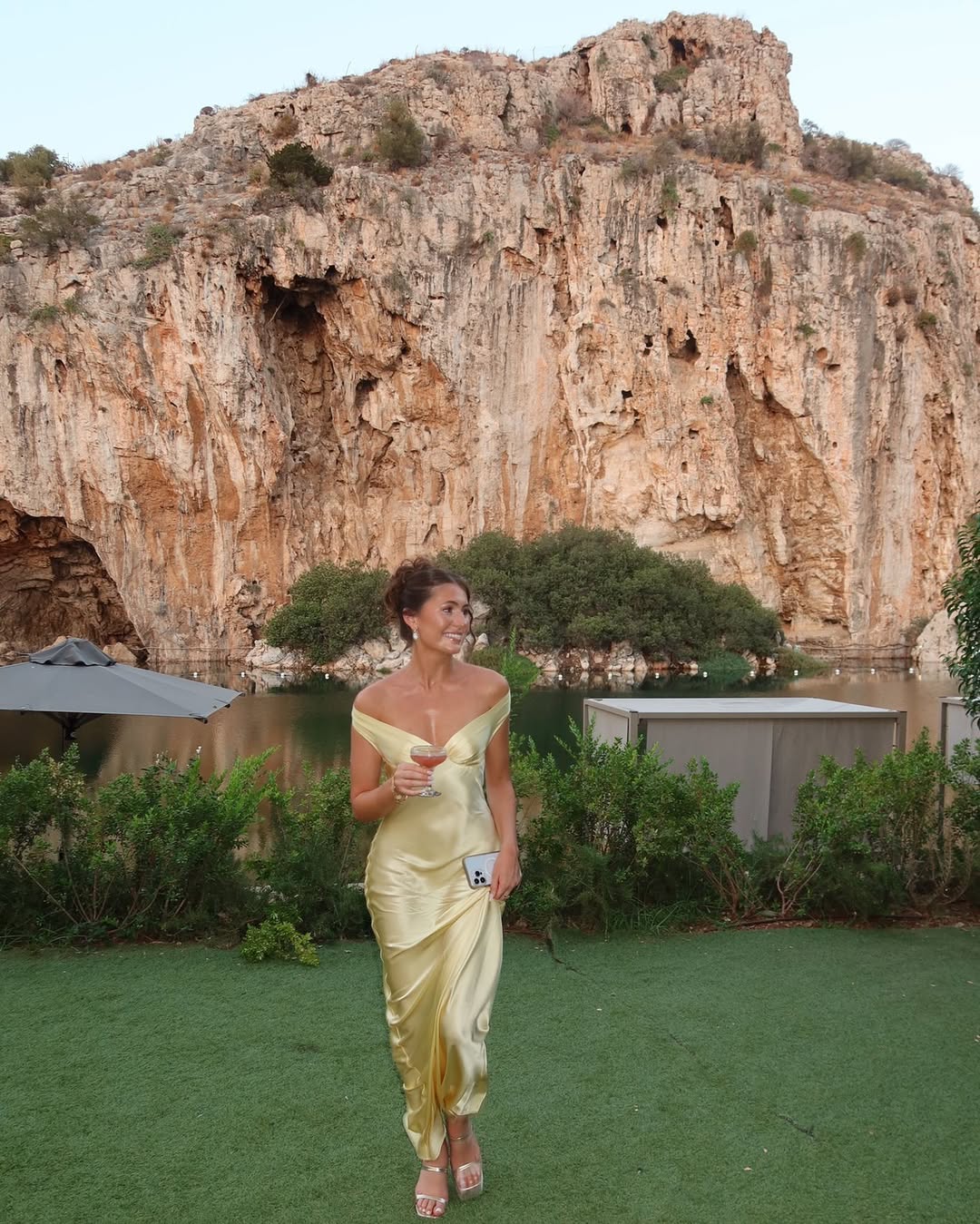 Elegant woman in a yellow satin off-shoulder maxi dress holding a champagne glass at a summer outdoor wedding near cliffs, perfect wedding guest look by Isadora London